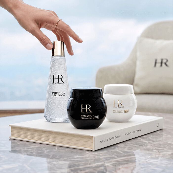 Drie huidverzorgingsproducten van Helena Rubinstein (Prodigy Cellglow en Re-Plasty) op een wit boek voor een raam met uitzicht op een landschap.​