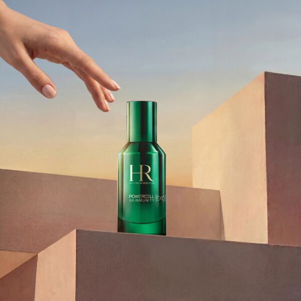 Productfoto van het Helena Rubinstein Powercell Skinmunity Exo Serum in een glanzende groene glazen fles. De fles staat bovenop een stapel roze geometrische podia, terwijl een elegante hand er van bovenaf naar reikt, tegen een warme, zonsondergangkleurige luchtachtergrond.​