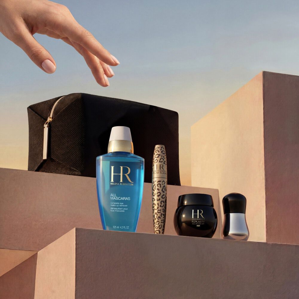 Een elegante hand reikt naar een zorgvuldig samengestelde selectie Helena Rubinstein producten, gerangschikt op minimalistische, zandbeige stenen blokken. De set wordt gepresenteerd tegen een warme, gouden zonsondergangshemel en bevat een zwart reisetui, de blauwe 'All Mascaras' oog-make-upremover, een Lash Queen Feline Blacks mascara met luipaardprint, een klein zwart Re-Plasty cremepotje en een zilveren applicator.​