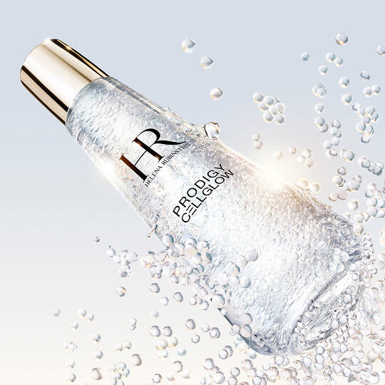 Helena Rubinstein Prodigy Cellglow serumfles gevuld met belletjes, gekanteld en omringd door sprankelende zwevende belletjes, op een lichte en heldere achtergrond.