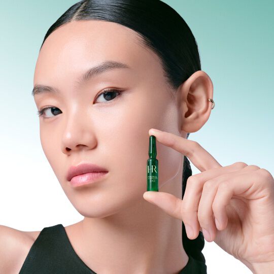 Close-up van het gezicht van een vrouw die teerkes een groene Powercell [EXO] Skinmunity ampul van Helena Rubinstein tussen haar vingers vasthoudt, tegen een groen-witte verloopachtergrond.​