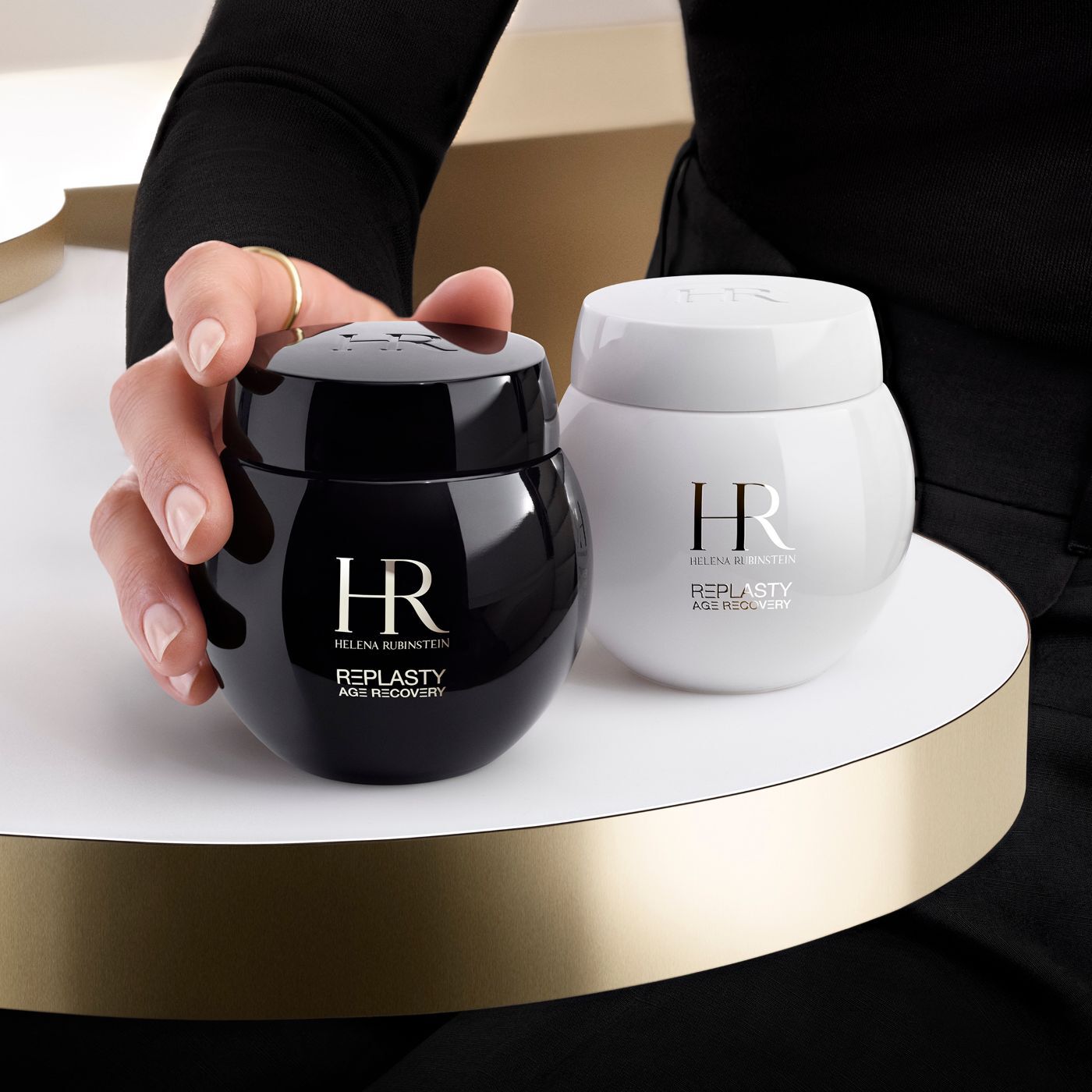 Een hand pakt een zwarte pot Helena Rubinstein Re-Plasty Age Recovery creme, met een bijpassende witte pot ernaast, op een gouden platform.