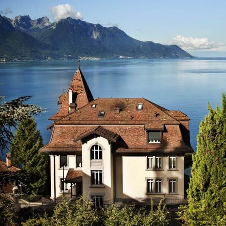 Een groot traditioneel huis met een rood pannendak en lichte muren, gelegen aan een groot diepblauw meer, omringd door bergen. De lucht is helder.