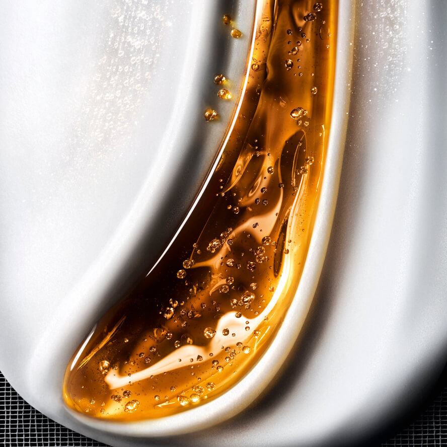 Close-up van een vloeibare cosmetische textuur, die golven van glanzende gouden gel met bubbels mengt op een cremekleurige witte achtergrond, wat een rijke huidverzorging oproept.