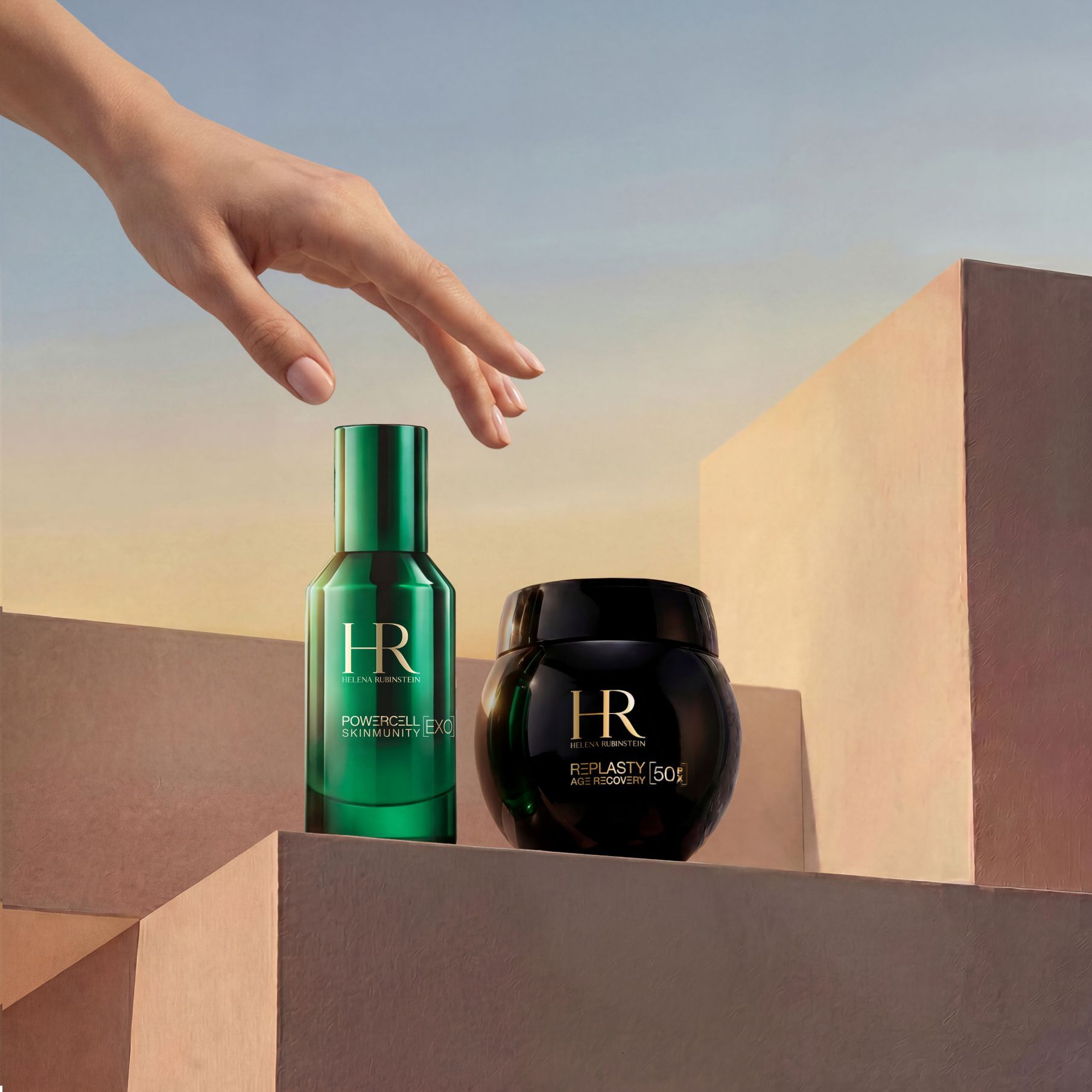 Een elegante hand die naar de Helena Rubinstein Duo Travel Replasty Night – Powercell Essence reikt, bestaande uit een groene fles Powercell Skinmunity Essence en een pot Re Plasty Age Recovery Night Cream, gepresenteerd op zanderige geometrische blokken tegen een warme zonsonderganghemel.​