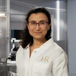 A scientist in a lab coat smiles confidently in a modern laboratory setting, with microscopes in the background.