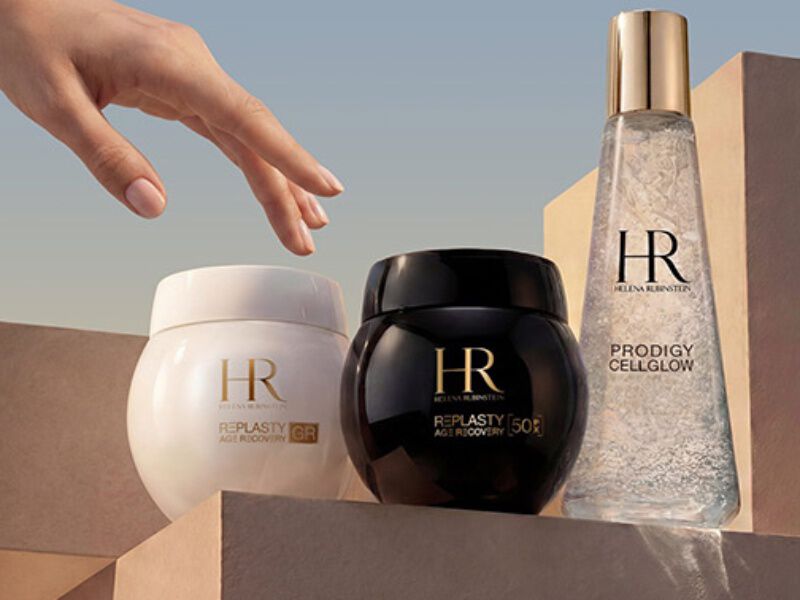 Een hand reikt uit naar drie producten van Helena Rubinstein, die een elegant ontwerp en zachte verlichting laten zien.