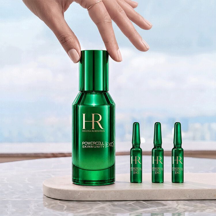 Een hand die de groene fles van het Helena Rubinstein Powercell [EXO] Skinmunity serum aanraakt, geplaatst naast drie [EXO] Shots ampullen op een marmeren standaard tegen de achtergrond van een helder landschap.​