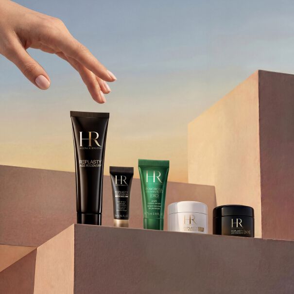 Productfoto van de maandelijkse selectie cadeaus van Helena Rubinstein. Onder een zacht verlopende hemel reikt een elegante hand naar luxe verzorgingsproducten op minimalistische zandkleurige blokken. Deze exclusieve set bevat een glazen flacon Powercell Skinmunity, een reistube Powercell Skin Rehab, twee reistubes Powercell Skinmunity Exo, een pot Re-Plasty Age Recovery Day Cream [GR] en een chic wit reisetui op de achtergrond.​