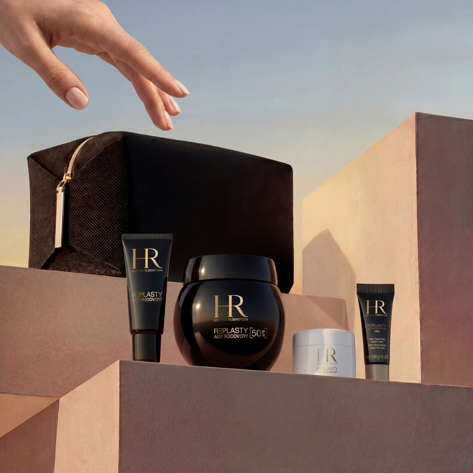 Een elegante hand reikt naar een Helena Rubinstein geschenkset, uitgestald op zandkleurige stenen blokken tegen een zonsonderganghemel, met een zwart limited edition reistasje en het Replasty Age Recovery assortiment: Eye Repairing Night Care, Day Cream, Night 50PX en Body Repairing Care.​