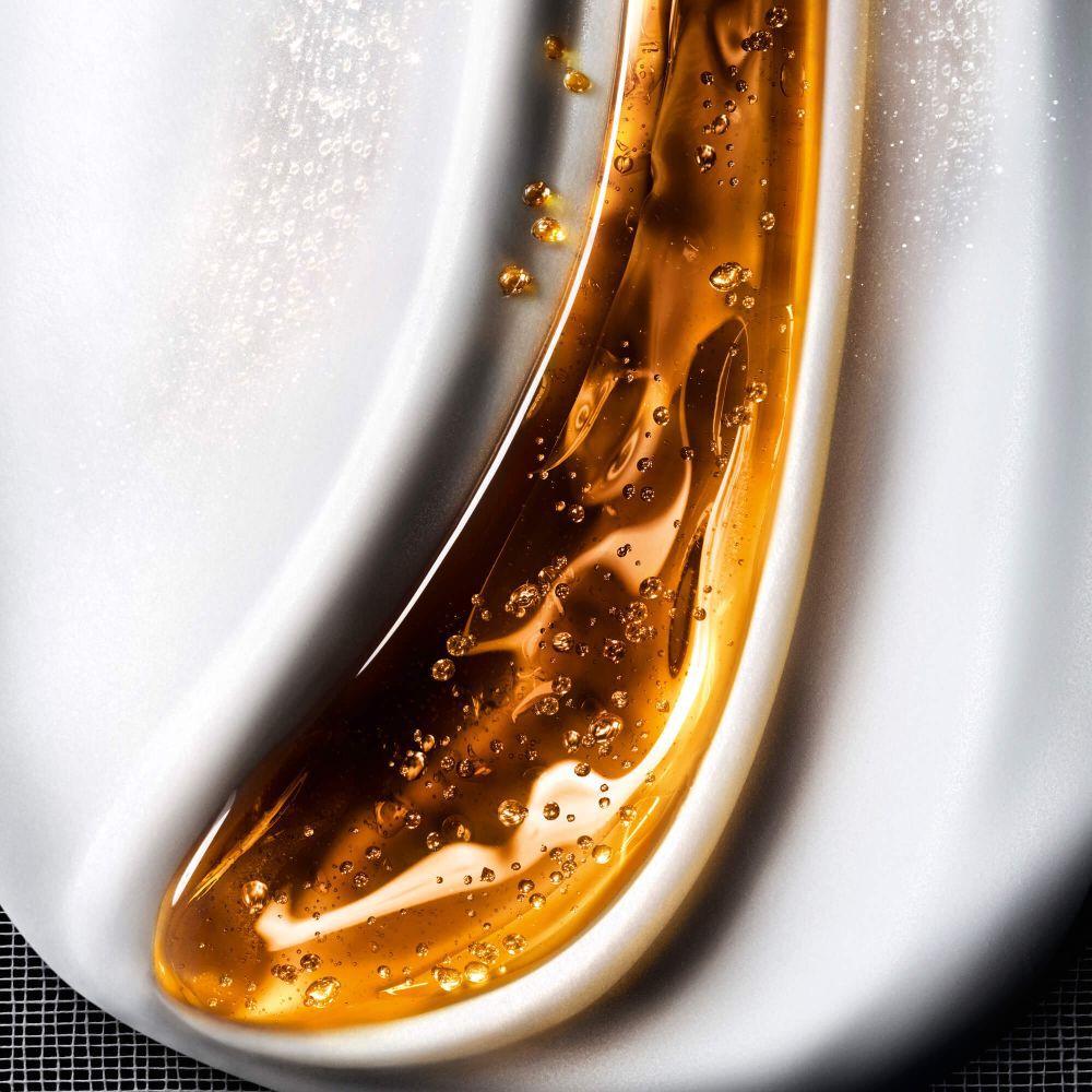  Close-up van een vloeibare cosmetische textuur, die golven van glanzende gouden gel met bubbels mengt op een cremekleurige witte achtergrond, wat een rijke huidverzorging oproept.